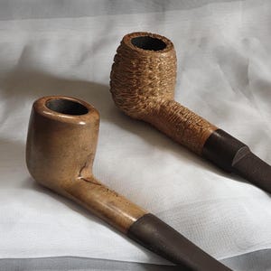May include: Two tobacco pipes are displayed. One is made of light brown wood with a dark brown stem. The other pipe is made of a textured, light brown material with a dark brown stem. Both pipes are resting on a white surface.