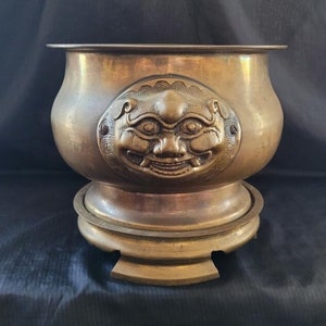 May include: A large, round, brass planter with a decorative lion's head design on the front. The planter has a wide base and a raised lip around the top.