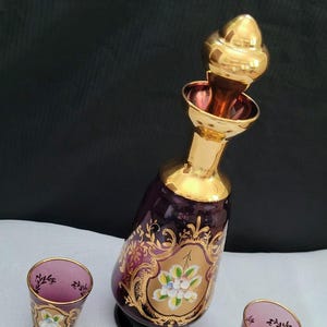 May include: A purple glass decanter with gold accents and floral designs, along with three matching shot glasses.