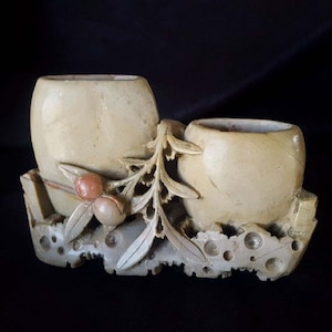 May include: A carved stone double vase with a leafy design and two small, round openings. The vase is a light beige color with brown and pink accents.
