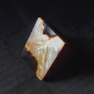 May include: A square-shaped ring with a polished, iridescent surface. The ring features a mix of brown, white, and cream colors, with a dark brown border. The ring is set against a dark background.