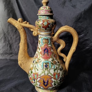 May include: An ornate ceramic pitcher with a gold-colored handle and spout. The pitcher is decorated with colorful floral patterns in pink, blue, and orange against a white background. The lid is topped with a gold finial. The spout is shaped like a dragon's head.