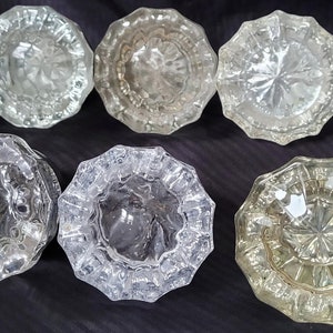 May include: Six clear glass door knobs with intricate patterns. The knobs have a variety of designs, including a starburst, a floral pattern, and a geometric design.