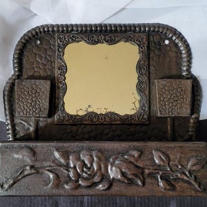May include: A vintage metal wall mounted mirror with two small compartments on either side of the mirror. The mirror has a decorative frame with a floral design. The bottom of the mirror has a decorative floral design.