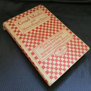 May include: A vintage cookbook titled "Good Meals and How to Prepare Them." The cover features a red and white checkered pattern with text in a serif font. The book is a guide to meal planning, cooking, and serving.