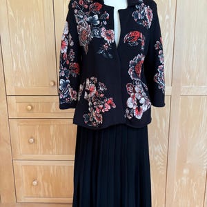 May include: A black jacket and skirt set. The jacket features a floral pattern in red, white, and pink. The skirt is a long, pleated design. The outfit is displayed on a mannequin against a light wood background.