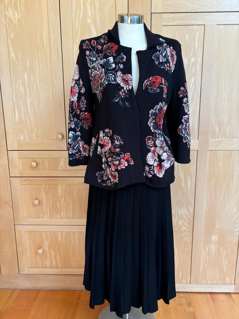 May include: A black jacket and skirt set. The jacket features a floral pattern in red, white, and pink. The skirt is a long, pleated design. The outfit is displayed on a mannequin against a light wood background.