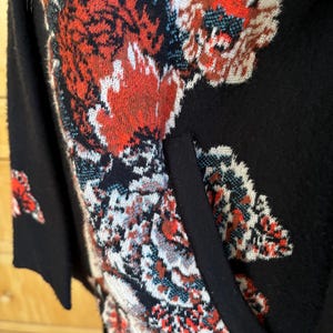 May include: Black cardigan with a floral pattern in red, white, and teal. The cardigan has a pocket and a black trim. The floral design is intricate and covers the front and sleeves of the garment. The cardigan is made of a soft material.