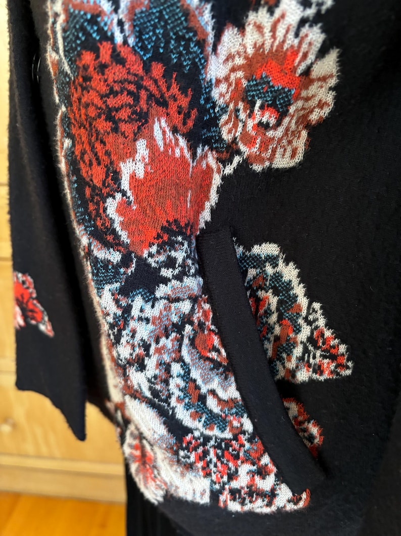 May include: Black cardigan with a floral pattern in red, white, and teal. The cardigan has a pocket and a black trim. The floral design is intricate and covers the front and sleeves of the garment. The cardigan is made of a soft material.