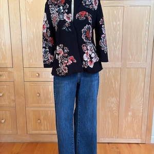 May include: A black floral blazer with red, white, and blue flower designs is paired with blue denim jeans. The blazer has a unique asymmetrical cut and is displayed on a mannequin.