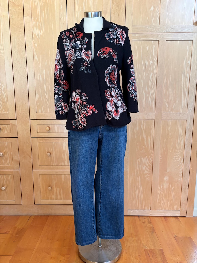 May include: A black floral blazer with red, white, and blue flower designs is paired with blue denim jeans. The blazer has a unique asymmetrical cut and is displayed on a mannequin.