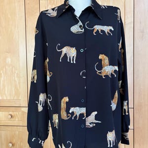 May include: Black button-down shirt with a leopard print design. The long-sleeved shirt features a classic collar and cuffs. The leopard print is in shades of tan, brown, and white, creating a striking contrast against the black fabric.
