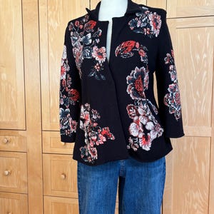 May include: A black jacket with a floral pattern in red, white, and gray. The jacket has a shawl collar and three-quarter sleeves. The jacket is paired with blue jeans.
