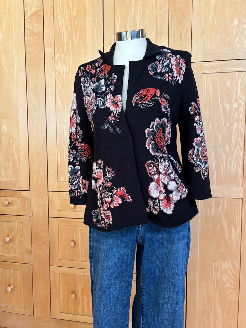 May include: A black jacket with a floral pattern in red, white, and gray. The jacket has a shawl collar and three-quarter sleeves. The jacket is paired with blue jeans.