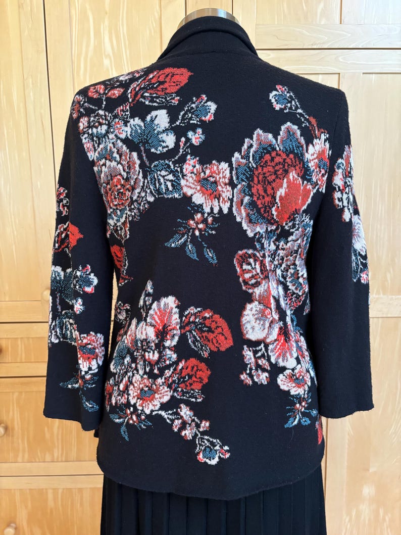 May include: Black jacket with a floral design in red, white, and blue. The jacket has a shawl collar and three-quarter sleeves. The floral pattern covers the back and sleeves of the jacket.