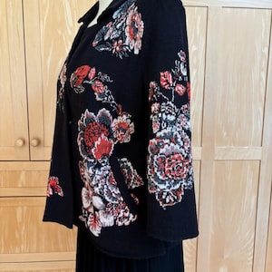 May include: A black jacket with a floral design in red, white, and gray. The jacket has a collar and is displayed on a mannequin. The floral pattern is concentrated on the sleeves and front of the jacket.