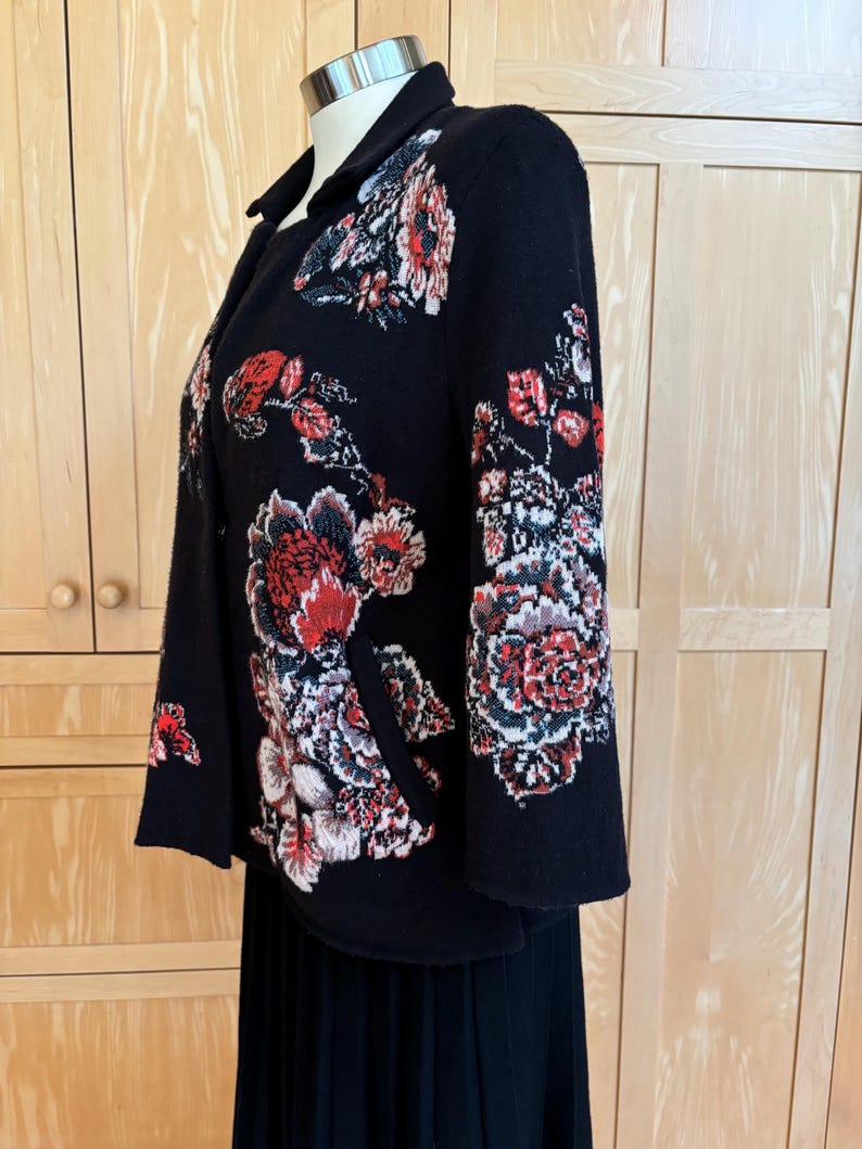 May include: A black jacket with a floral design in red, white, and gray. The jacket has a collar and is displayed on a mannequin. The floral pattern is concentrated on the sleeves and front of the jacket.