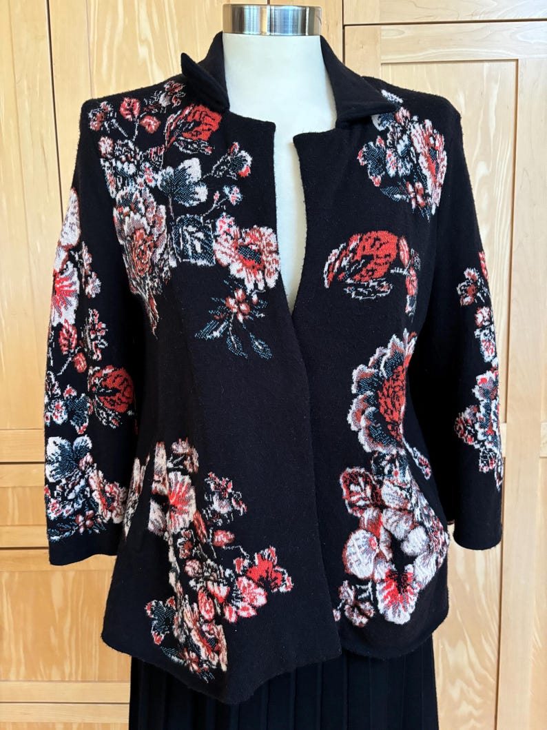 May include: Black open-front jacket with a floral pattern in red, white, and gray. The jacket has three-quarter sleeves and a collar. The floral design is concentrated on the shoulders, sleeves, and lower front panels.
