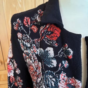 May include: Black cardigan with a floral pattern in red, white, and blue. The cardigan has a collar and is made of a soft, textured material. The floral design is intricate and covers the front and shoulders of the garment.