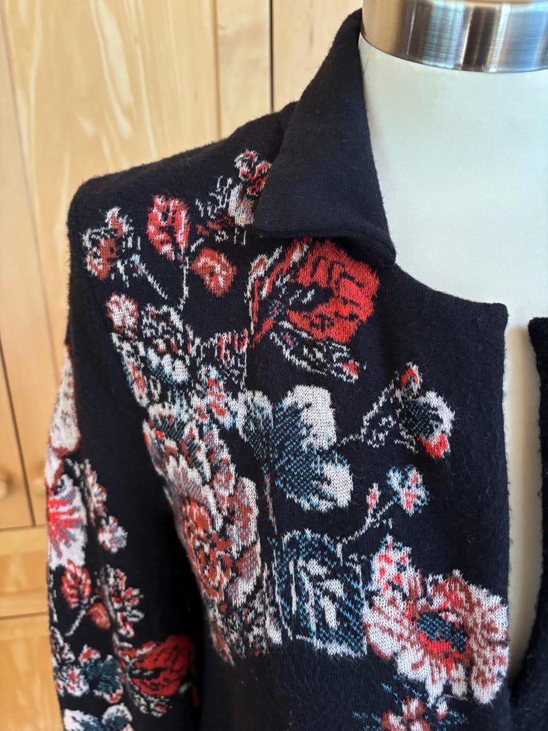 May include: Black cardigan with a floral pattern in red, white, and blue. The cardigan has a collar and is made of a soft, textured material. The floral design is intricate and covers the front and shoulders of the garment.