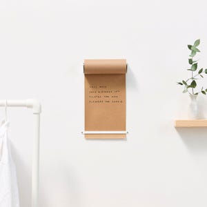 Op de afbeelding: Een bruine papierrol met een wit metalen frame is gemonteerd op een witte muur. Het papier heeft handgeschreven notities die zeggen "Call Rose", "Jack Birthday 10th", "Pilates 5pm Wed" en "Flowers for Sophie".