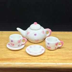 May include: A miniature white ceramic tea set with pink heart and purple dot designs. The set includes a teapot, two cups, and two saucers. The teapot has a pink handle and a pink knob on the lid.