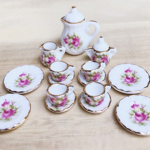 May include: A miniature porcelain tea set with a floral rose design. The set includes a teapot, creamer, sugar bowl, cups, saucers, and plates. Each piece is white with pink roses and gold trim, creating a delicate and elegant aesthetic.