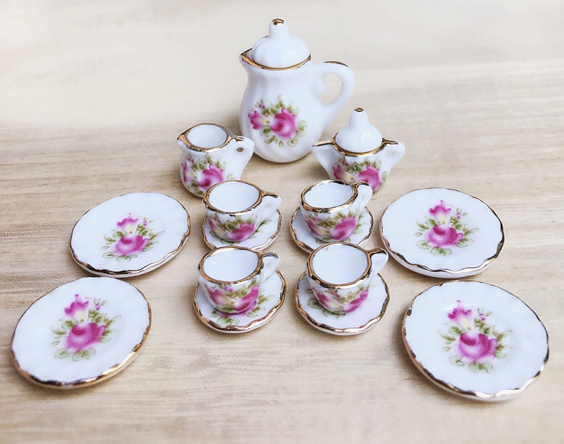 May include: A miniature porcelain tea set with a floral rose design. The set includes a teapot, creamer, sugar bowl, cups, saucers, and plates. Each piece is white with pink roses and gold trim, creating a delicate and elegant aesthetic.