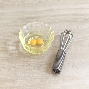 Peut inclure: Une sc&egrave;ne de cuisine miniature avec un petit bol jaune &agrave; bords festonn&eacute;s contenant deux jaunes d'&oelig;ufs et un fouet argent&eacute;. Le fouet a une poign&eacute;e en spirale. Les articles sont pos&eacute;s sur une surface de couleur claire.