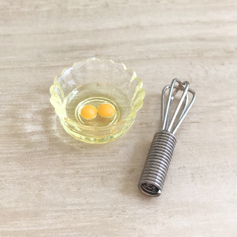 Peut inclure: Une sc&egrave;ne de cuisine miniature avec un petit bol jaune &agrave; bords festonn&eacute;s contenant deux jaunes d'&oelig;ufs et un fouet argent&eacute;. Le fouet a une poign&eacute;e en spirale. Les articles sont pos&eacute;s sur une surface de couleur claire.