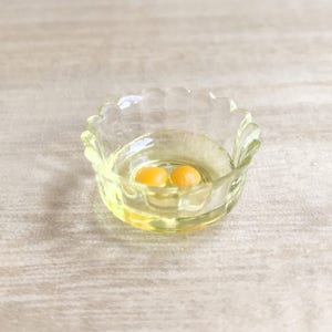 Op de afbeelding: Een kleine, heldere glazen kom met een geschulpte rand, met twee eierdooiers in een vloeistof. De kom heeft een lichte gele tint en staat op een licht oppervlak. De dooiers zijn fel oranje.