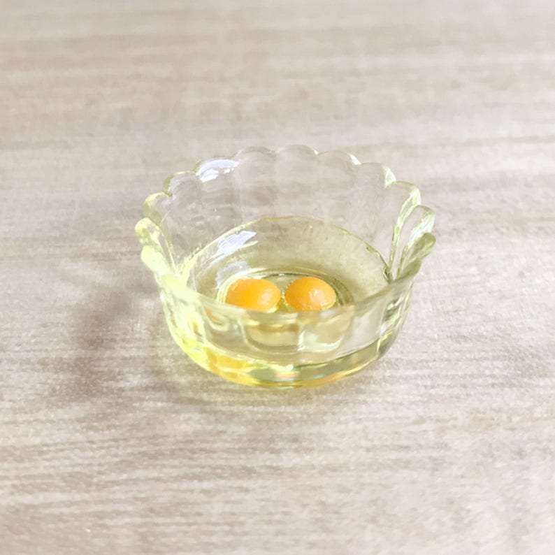 Op de afbeelding: Een kleine, heldere glazen kom met een geschulpte rand, met twee eierdooiers in een vloeistof. De kom heeft een lichte gele tint en staat op een licht oppervlak. De dooiers zijn fel oranje.