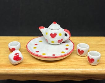 May include: Miniature ceramic tea set with a white base, red heart and yellow dot accents. The set includes a teapot, a plate, and three cups. The teapot has a red handle and spout. The plate and cups have red heart designs.