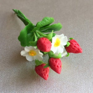 May include: A bouquet of five red strawberries and three white flowers with yellow centers. The strawberries and flowers are made of polymer clay and are attached to green stems.