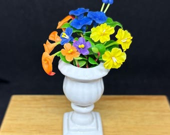 Puede incluir: Un pequeño jarrón de cerámica blanca lleno de flores coloridas hechas a mano. El arreglo incluye flores azules, amarillas, naranjas y moradas con hojas verdes. El jarrón está sobre una superficie de madera contra un fondo oscuro, mostrando el diseño floral detallado.