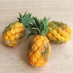 Op de afbeelding: Drie miniatuur ananassculpturen. De ananassen zijn geel en groen, met gedetailleerde texturen en groene, bladachtige toppen. De sculpturen zijn gerangschikt op een lichtgekleurd oppervlak, wat hun ingewikkelde ontwerp laat zien.
