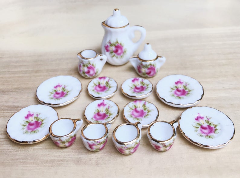 May include: Miniature porcelain tea set with a floral rose design. The set includes a teapot, creamer, sugar bowl, plates, and cups. Each piece is white with pink roses and gold trim, perfect for dollhouses or display.