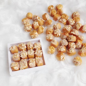 May include: A white box filled with miniature pastries topped with almonds and sugar. The pastries are arranged in a grid pattern. There are also loose pastries scattered on a white fluffy surface.