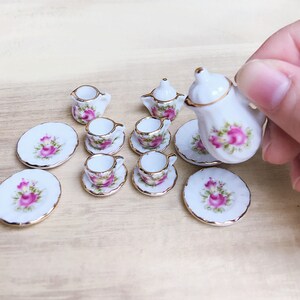 May include: Miniature porcelain tea set with a floral rose design. The set includes a teapot, creamer, sugar bowl, cups, saucers, and plates. Each piece is white with pink roses, green leaves, and gold trim. The set is designed for a dollhouse.
