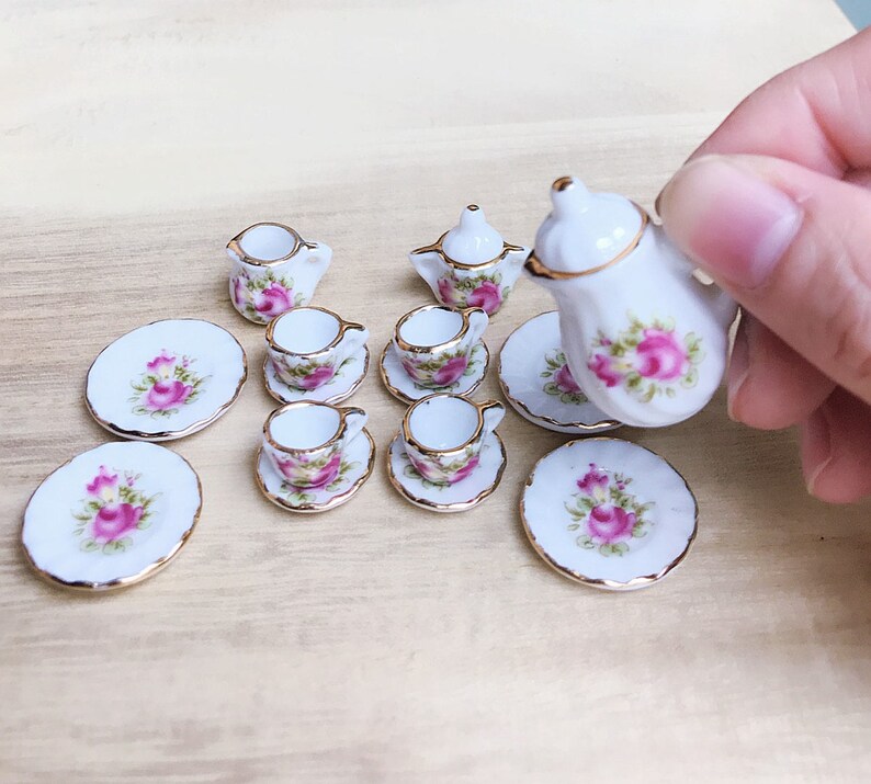 May include: Miniature porcelain tea set with a floral rose design. The set includes a teapot, creamer, sugar bowl, cups, saucers, and plates. Each piece is white with pink roses, green leaves, and gold trim. The set is designed for a dollhouse.