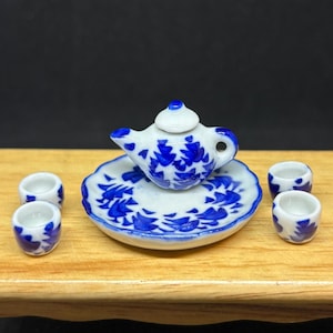 May include: Miniature white and blue porcelain tea set. The set includes a teapot, a saucer, and three small teacups. The teapot and saucer feature a blue floral design. The teacups have a similar blue pattern.