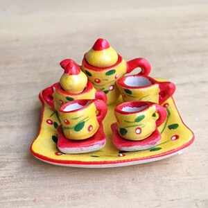 May include: A miniature tea set with a yellow teapot, creamer, sugar bowl, and four cups and saucers. The set is decorated with red and green floral designs and is displayed on a yellow tray with a red rim.