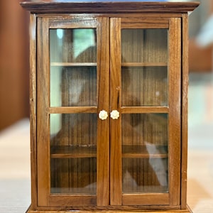 Puede incluir: Un pequeño armario de madera con dos puertas de cristal. El armario es de color marrón cálido con una parte superior e inferior marrón oscuro. Las puertas tienen pequeños pomos blancos en forma de flor. En el interior se ven varias estanterías.