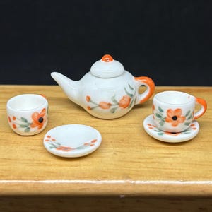May include: Miniature white porcelain tea set with orange floral accents. The set includes a teapot, two cups, and a saucer. The teapot has an orange handle and spout. The cups and saucer have orange flower designs and green leaves.