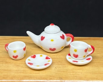 May include: A miniature white ceramic tea set with red heart and yellow dot designs. The set includes a teapot, two cups, and two saucers. The teapot has a red handle and a red top. The cups and saucers have red heart and yellow dot designs.