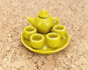 May include: A miniature chartreuse tea set consisting of a small teapot with a lid and four matching handleless cups arranged on a round, slightly wavy-edged saucer. The entire set features a smooth, glossy finish and a uniform bright yellow-green color.