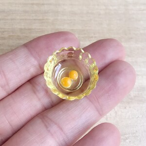 Op de afbeelding: Een miniatuur, heldere gele kom met een geschulpte rand, met daarin twee kleine, ronde, oranje dooiers. De kom staat tegen een neutrale achtergrond. Dit item is waarschijnlijk een poppenhuisaccessoire of een miniatuurvoedselartikel.