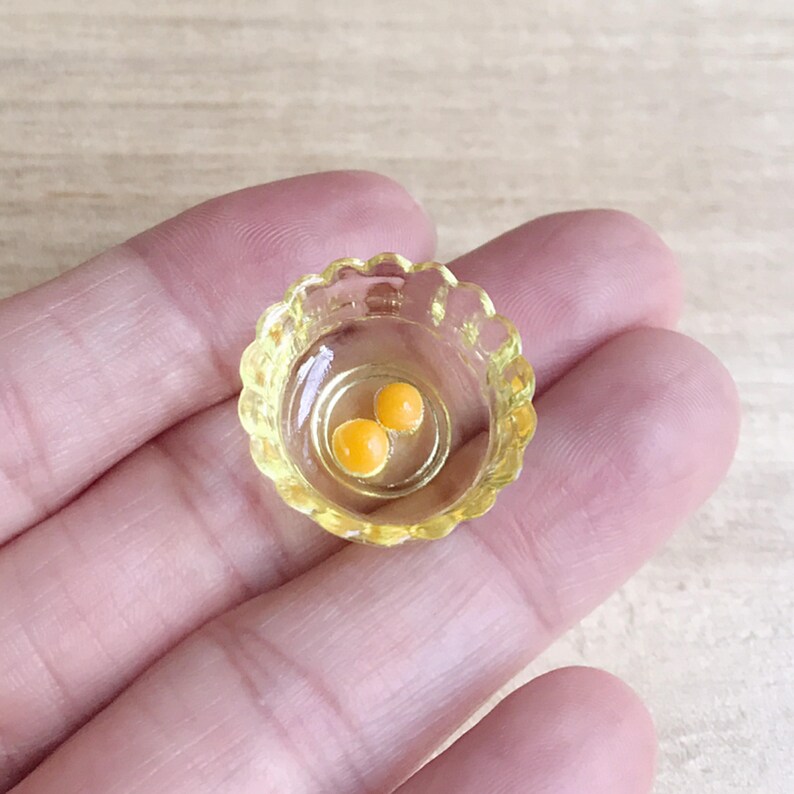 Op de afbeelding: Een miniatuur, heldere gele kom met een geschulpte rand, met daarin twee kleine, ronde, oranje dooiers. De kom staat tegen een neutrale achtergrond. Dit item is waarschijnlijk een poppenhuisaccessoire of een miniatuurvoedselartikel.