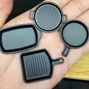 May include: Four black miniature cast iron frying pans, two with ridged surfaces and two with smooth surfaces. The pans are arranged on a hand.