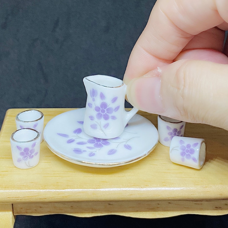 May include: A miniature tea set with a white pitcher and four cups, all decorated with a purple floral pattern. The set is on a wooden table.